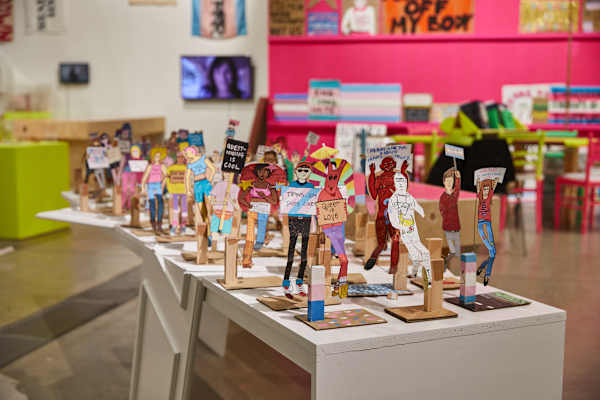 An image of a gallery with a pink wall displaying protest placards in the background. In the foreground and in focus, a table of human-shaped paper models in a variety of different styles and clothing hold celebratory and protest signs, including messages 'Queer is love' and 'Trans joy saves lives'