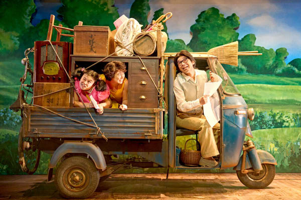 Image shows a tuk-tuk with household items on top. There is a man in the front of the vehicle with a newspaper in his hand and he is speaking to two actors playing young children who are sticking their heads out of the window