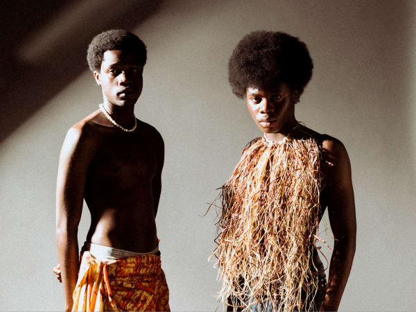Two people standing facing the camera. One is topless and wearing a beaded/pearl necklace, with a yellow and orange dyed piece of fabric wrapped around as a skirt. The other is wearing a grass-like halterneck top. 