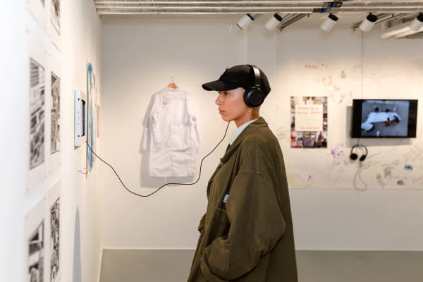 A person wearing a brown jacket and black cap stands, side profile to the camera, with headphones on facing a wall looking at artwork. In the background, more artworks can be seen hanging on the walls.