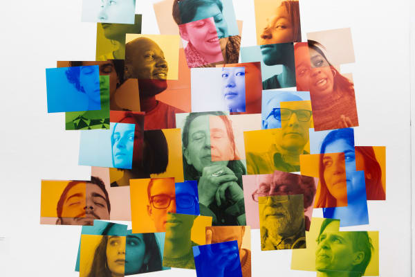 A vibrant photo collage featuring overlapping portraits of individuals, with each face tinted in bright colors like blue, orange, yellow, and green. The layered arrangement creates an abstract, mosaic-like effect, blending expressions and perspectives in a visually dynamic way