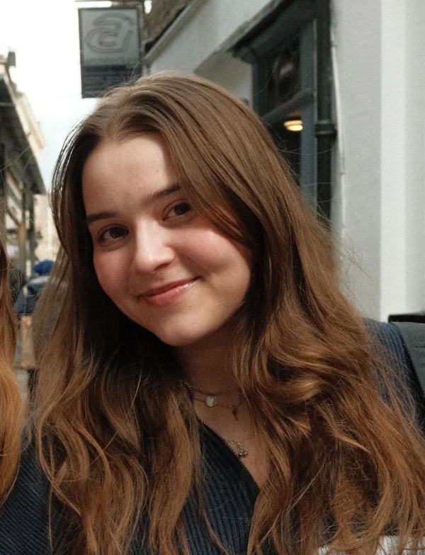 Headshot of Natalia Bednarova. Young woman with long brown hair and a subtle smile stands outdoors on a city street, dressed in a dark top. 