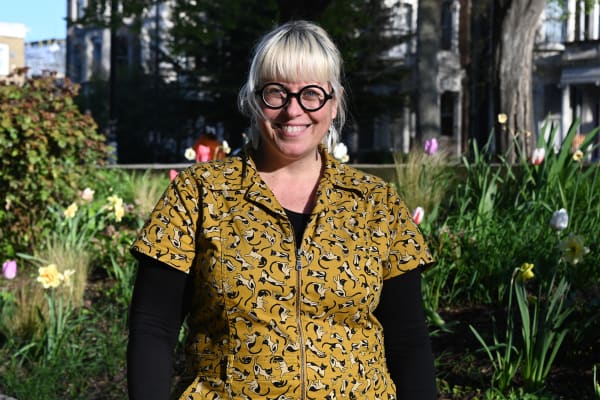 Image shows a blonde woman from waist up. She is smiling and is wearing thick black frame circle glasses, a yellow boilersuit with black cats on it, and a black under-shirt. In the background, there are tulips and other flowers.
