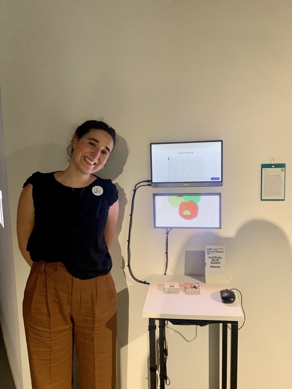 Smiling woman in black shirt and brown pants stands beside a tech display with screens showing text and graphics. 