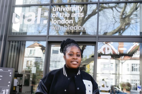 Image shows a young person standing outside the main entrance to Wimbledon College of Arts, UAL. The person is smiling against the backdrop of the campus, mostly made of glass.
