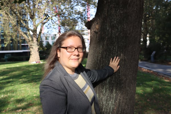 A white woman wearing glasses. She has mid-length hair and is wearing a grey suit jacket and striped t-shirt. She is touching a tree behind her and facing us.
