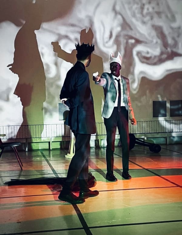 a photograph of a scene in BA Theatre Design student's Macbeth interpretation. Two characters are shown, one is pointing a gun at the other. Their silhouettes are shown on the wall behind them. 