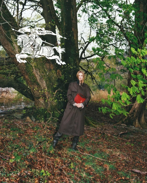 Burberry 2024 campaign: a model standing in woodland surroundings, wearing a brown trench coat, black boots and holding a red bag. Photography by Tyrone Lebon.