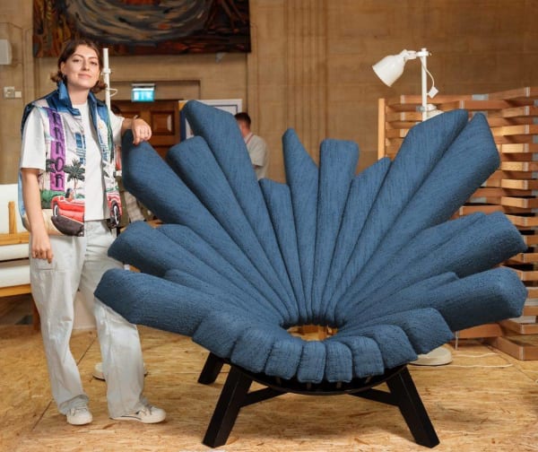 Maria Gill stands wearing a colourful vest, trousers, and trainers. She is leaning on her blue ‘All Seeing Chair’ which is positioned next to her. 