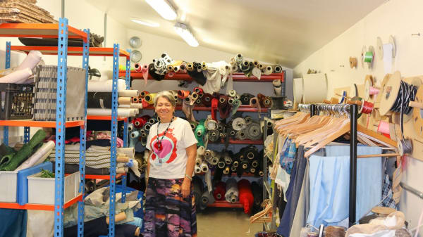 A white woman with short hair is standing in a store with lots of fabric rolls on shelves behind her and on each side. She is wearing a t-shirt with a printed pattern, a necklace and a skirt. 