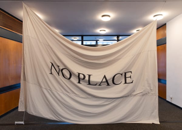 Large piece of material strung from the ceiling with 'NO PLACE' printed in a large black font in the middle.