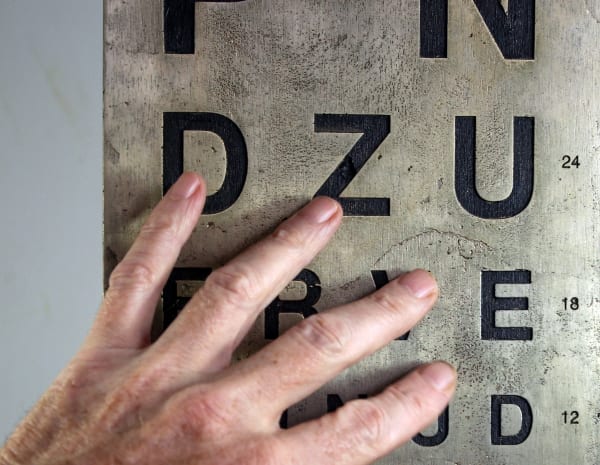 The image shows a splayed left hand touching a bronze plate upon which are lines of single black letters that are recessed into the plate. The letters are from a Snellen eye test chart and reduce in size with the larger letters being at the top and the smaller at the bottom.