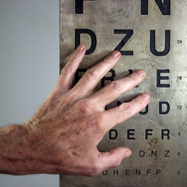 The image shows a splayed left hand  touching a bronze plate upon which are lines of single black letters that are recessed into the plate. The letters are from a Snellen eye test chart and reduce in size with the larger letters being at the top and the smaller at the bottom.