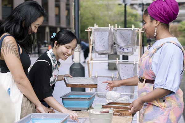 Two East Asian women with dark hair standing across a table from a south Asian woman wearing a headscarf and colourful dungarees are making paper using mesh screens. Handmade paper is on a drying rack in the background.