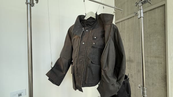 Dark gray jacket with a high collar and multiple buttons hangs on a minimalist metal display rack. The setting is modern, with a concrete wall.