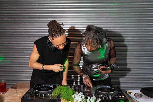 Omi and Linett standing behind the DJ decks, looking down 
