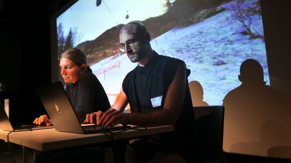 Lia Mazzari and Samuel Hertz performance sat at laptops with projected image of snowy landscape in background