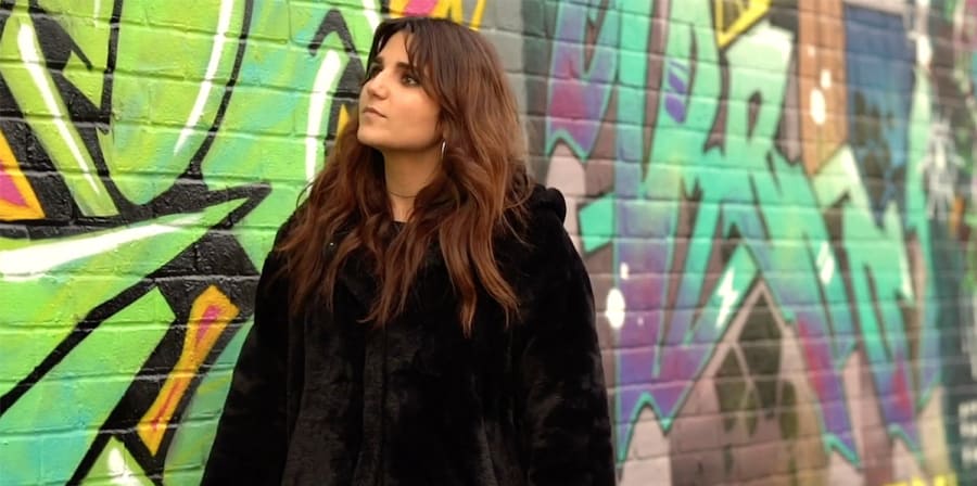 Revital stands in front of a heavily graffiti-ed brick wall.