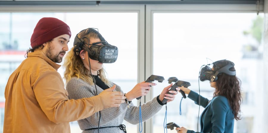 A man in a red hat helps a woman in a grey cardigan with a virtual reality headset. In the background is a woman also using virtual reality technology.