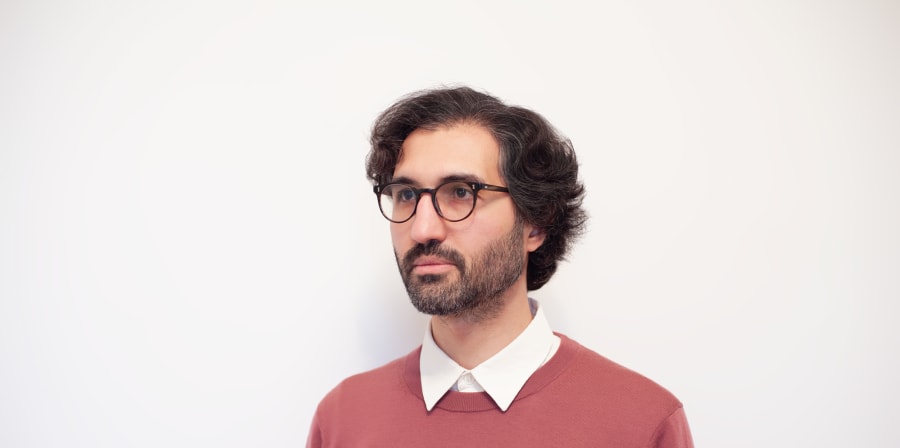 Head and chest of a man with dark hair and glasses wearing a white shirt and teracotta coloured jumper
