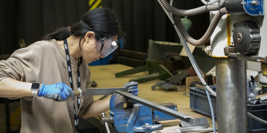 A student in gloves and protective head gear is working with metal equipment in the workshop
