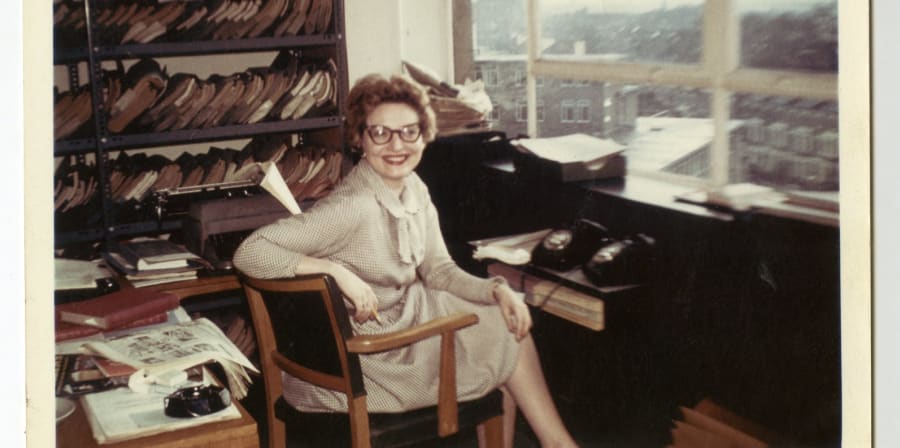 A light-haired woman wearing glasses and a beige dress sits smiling at a desk in front of a window and shelf of documents.