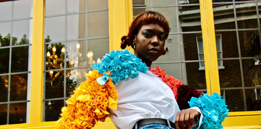  A lady wearing a vibrant coloured and ruffled shirt stands in front of a bright yellow building.