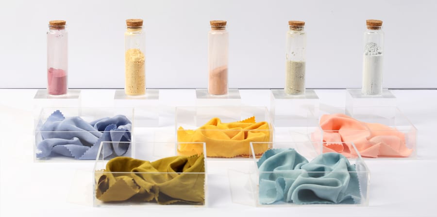 5 corked tubes with coloured dye powders within them, placed behind boxes with different coloured fabrics in, reflecting the dye pigments. 