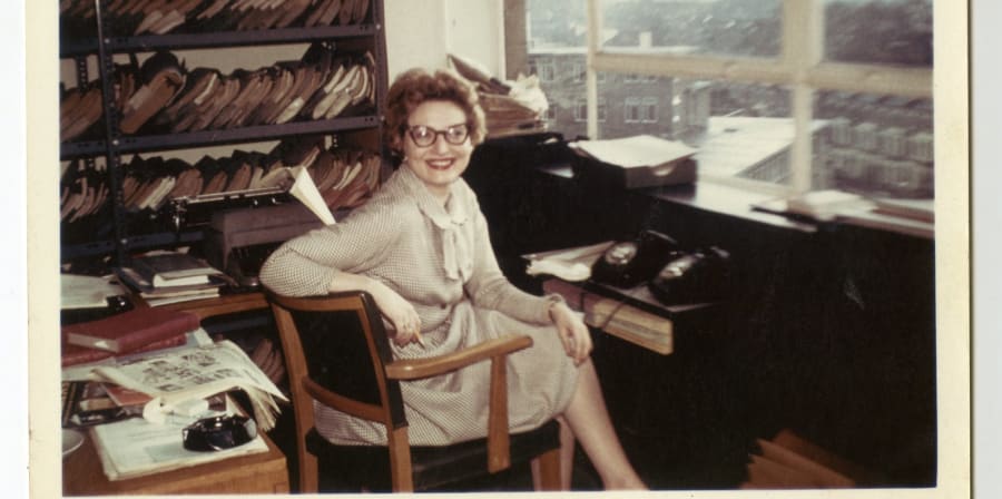 Archival photograph of a woman, Betty Willingale, sitting in an office, smiling, with shelves of paper script documents in the background