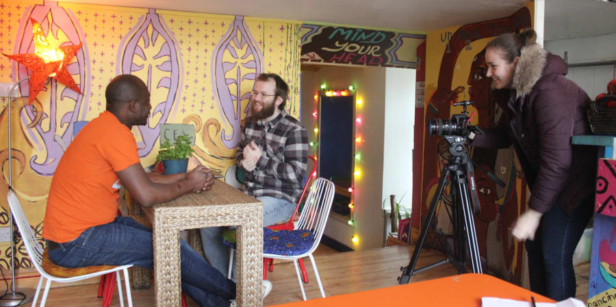 Two men, one client and one student, sit on a table in a cafe while a camera-woman films