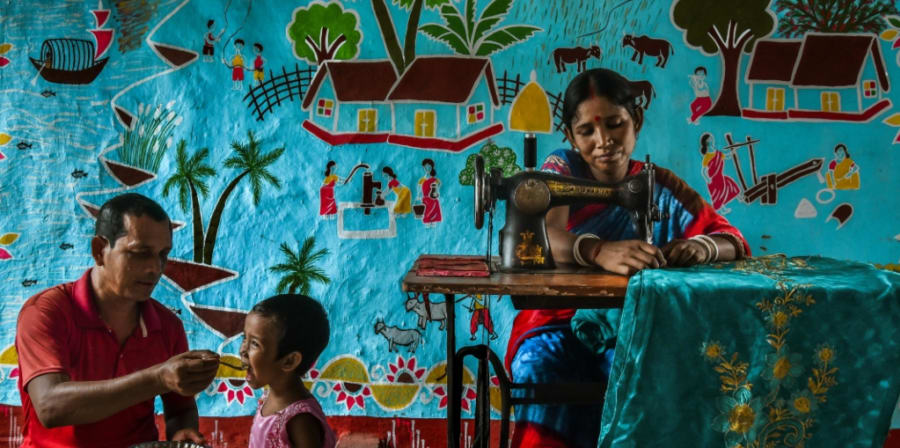 Lady at a sewing machine creating bright blue garment. Father and son sit nearby.