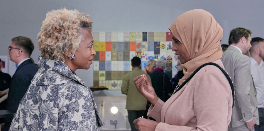 Two women of colour speaking in the foreground with a patchwork image display in the background