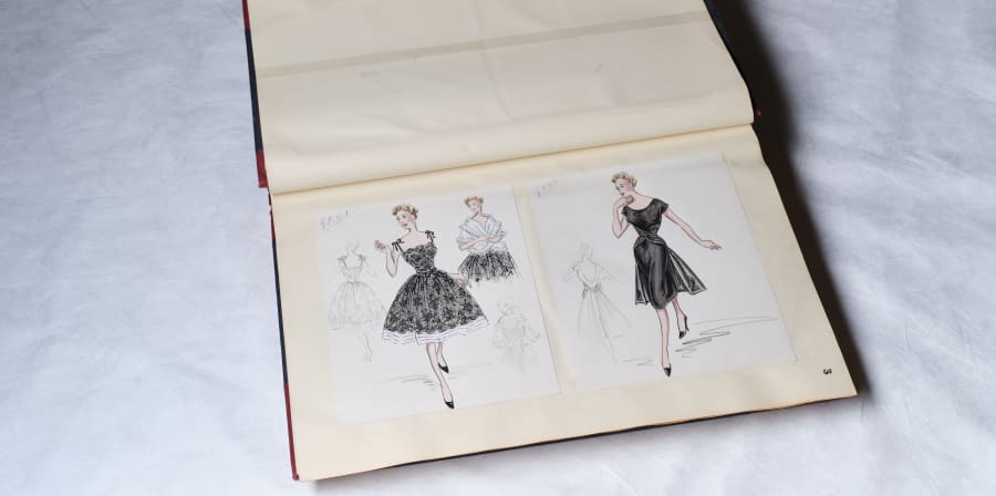 An open A3 sketchbook sits against a white backdrop. The pages feature two fashion sketches of blonde women, both wearing black dresses. One of the dresses is a lace with a cinched waist, and full circle skirt. The other is black and silky, with a cinched waist and flowy midi skirt.
