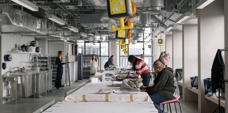 Long table with machinery on it. People sitting and standing at sides of table working.
