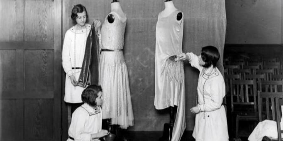 A black and white photo of three students dressing mannequins with dresses, students are wearing white coats and have short 1920s-style bob haircuts, two mannequins featuring 1920s-style dresses.