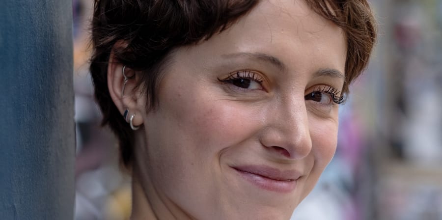 Smiling woman with short hair leaning against a wall in an urban setting.