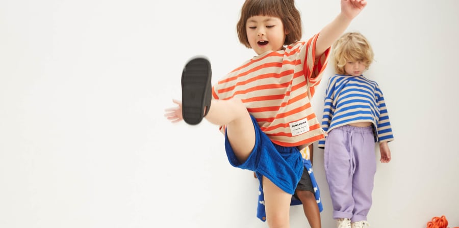 2 children, one standing at the back of a room against a wall and one playing and kicking their leg in the air.