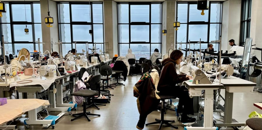 Room with sewing machines, with backdrop of glass windows. Person working on a machine near the front of the image.