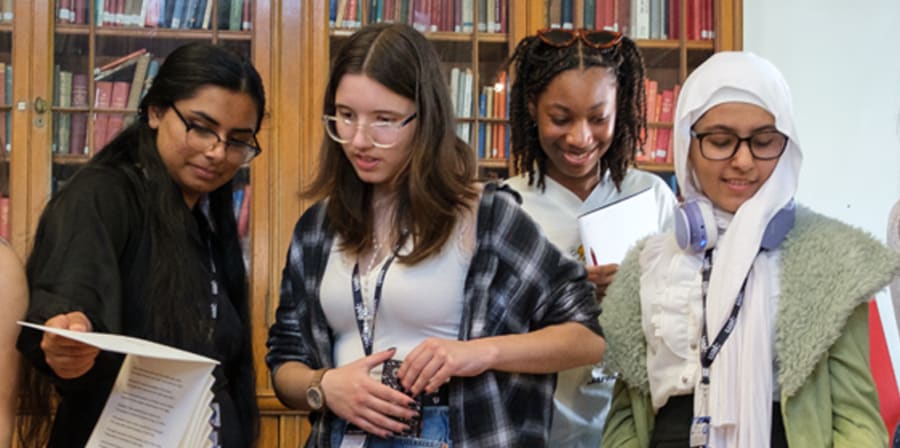 A group of students stand together in the library looking at books and objects