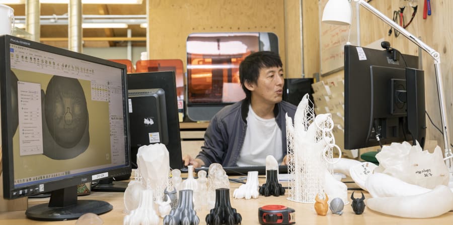 A student seated at a table with a monitor, lamp and lots of small models and prototypes of different shapes and sizes.