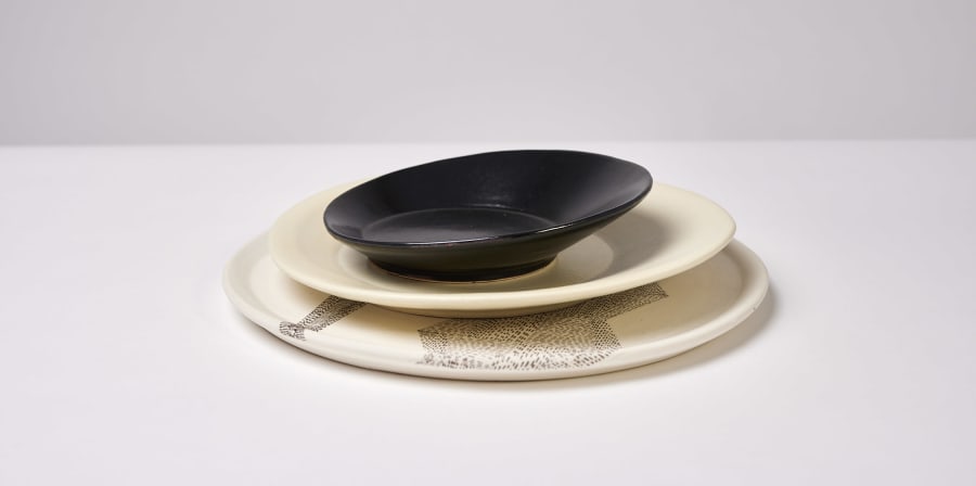 A small, black, slanted plate sits on top of 2 increasingly larger beige plates on a white tablecloth