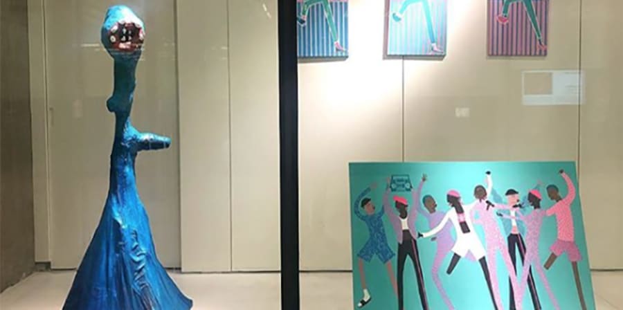 Photograph of a blue sculpture and paintings of illustrated people dancing on display in the Window Gallery at Central Saint Martins