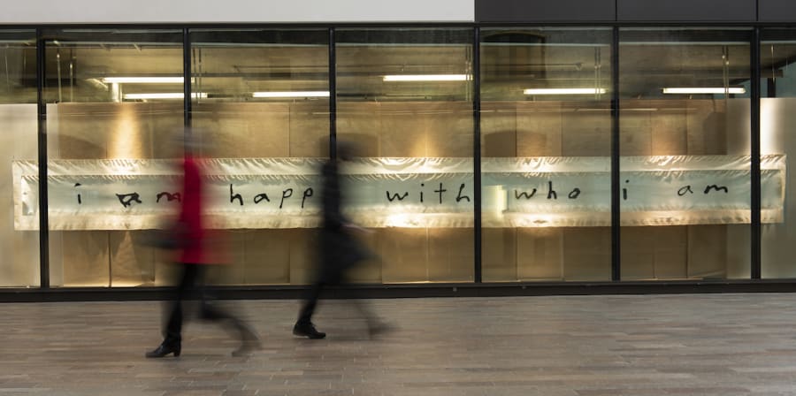 people are blurred walking past a long window, inside the window is a long fabric hanging that reads 'i am happy with who i am'