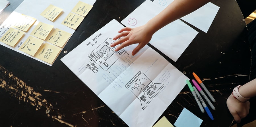 A student draws concepts on a desk filled with paper and post-it notes.