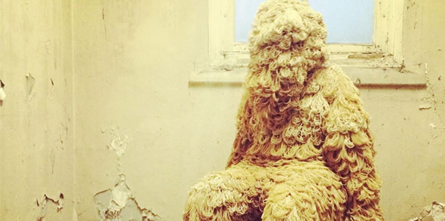 Image of a wooly human figure sat on a chair in a dilapidated room