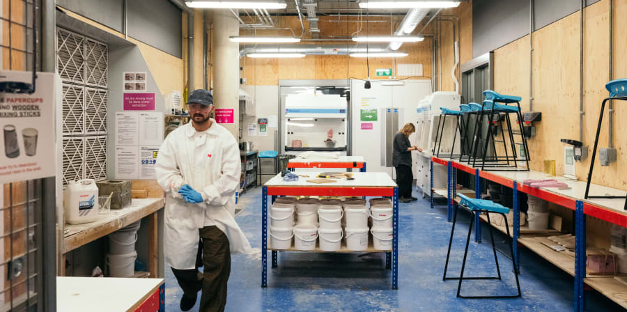 A wide shot of the casting workshop with a central island storing white buckets and a long-tabletop. At the far end a student is working at a large machine. Toward the left of the image, a technician in white lab coat, blue gloves and safety goggles is walking toward the camera.