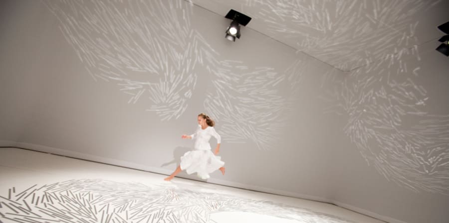 A person running around a room in a white dress. The floor of the room is covered in white marks. 
