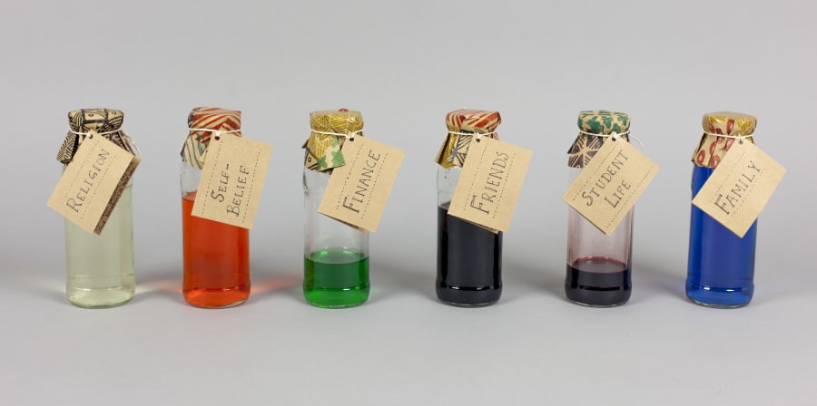 Six clear glass bottles with cardboard labels filled with coloured liquid 