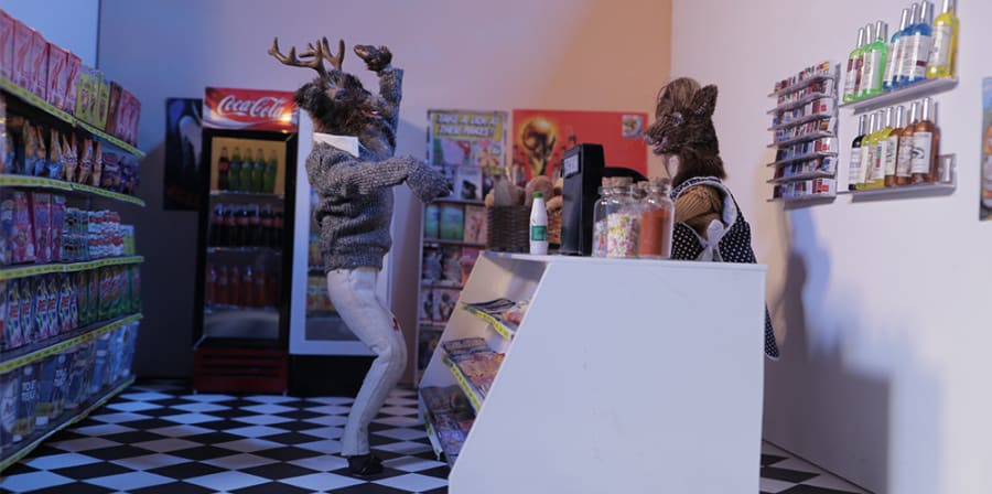 A male model of a deer, taken as a still from a stop motion animated film, waves to another deer in a shop scene.