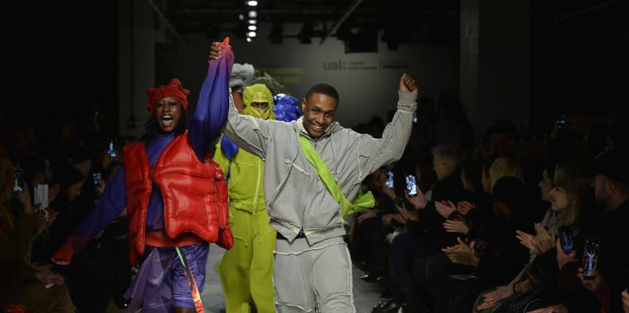 Yaku Stapleton and one of his models walks up the MA Fashion catwalk. He wears grey garments and wears a yellow over-body bag. The model next to him is wearing clothes made from materials that make it look like a puffer jacket. The garments she wears are bright red and purple. Both are smiling broadly. 